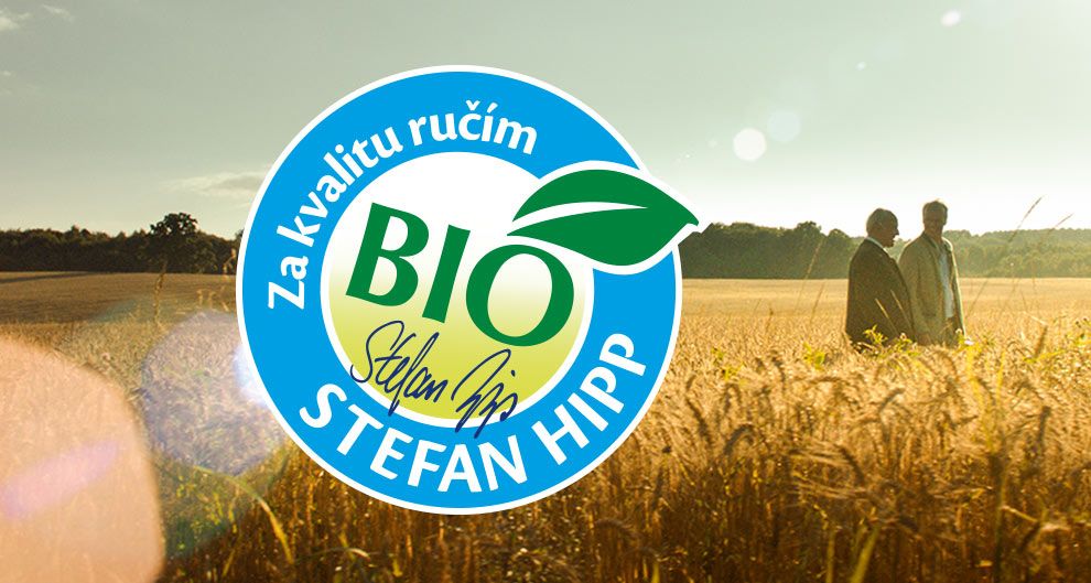 Dva muži stojí na pšeničném poli pod jasnou oblohou a na obrázku je velké modrobílé kruhové logo.