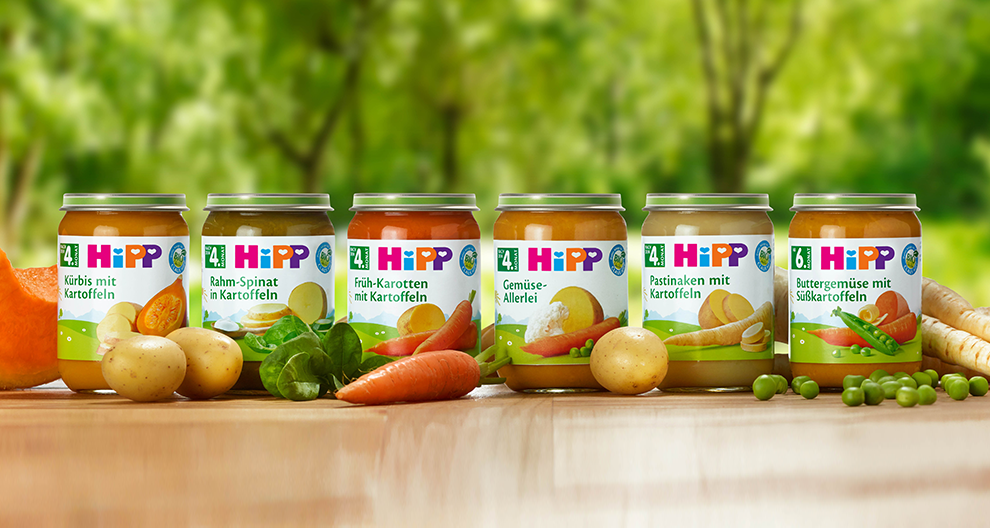 Jars of baby food with various labels are displayed on a wooden surface, surrounded by raw vegetables.