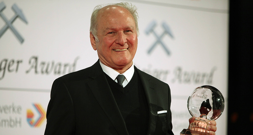 Starší muž v čiernom obleku stojí na pódiu a drží priehľadnú guľovú cenu so siluetou osoby na pozadí s nápisom "Schweizer Award" a logom a slovami "Schweizer Kreditbahn".